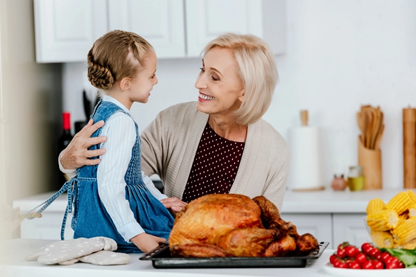 Little girl sits on kitchen counter beside cooked turkey as grandma holds her back.