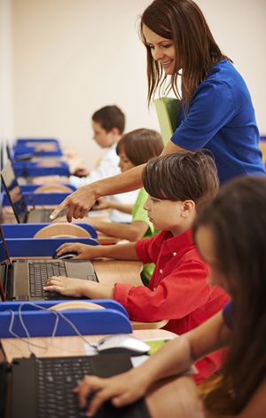 Row of kids leaning on computers with teacher giving individual instruction.