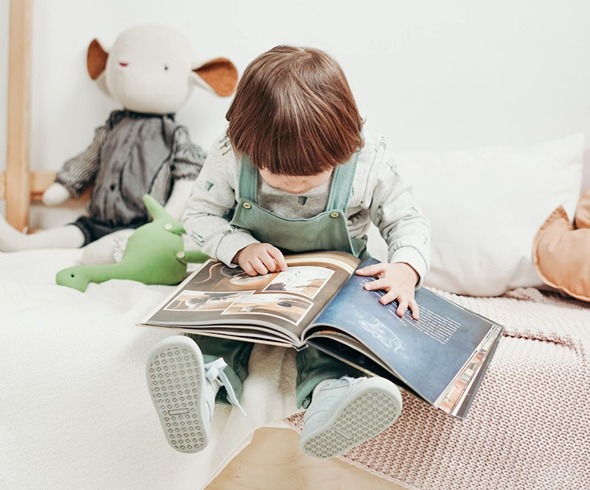 Toddler sitting on the edit of his bed looking at a picture book.