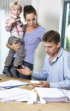 Woman holding daughter while looking over her partner who is working on the family budget at kitchen table.