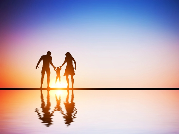 Silhouette of two parents holding child up child between them as the sun goes down.