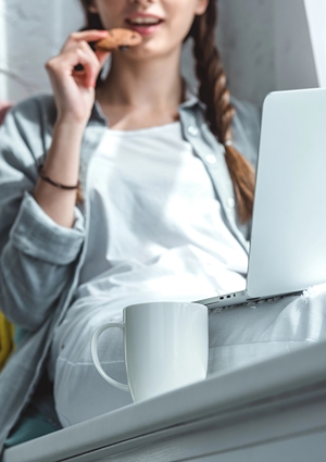 Teen girl eating a cookie with a coffee while using laptop