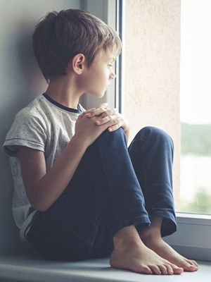 Sad boy seated on window ledge looking outside.