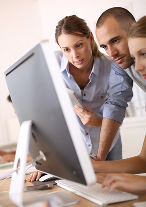 Two women and a man standing over desk while one of them is typing on computer.