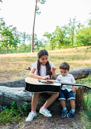 Sister and brother playing with a guitar while sitting on a log outdoors.