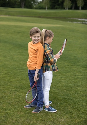 Girl and Boy standing back to back on grass holding tennis rackets.