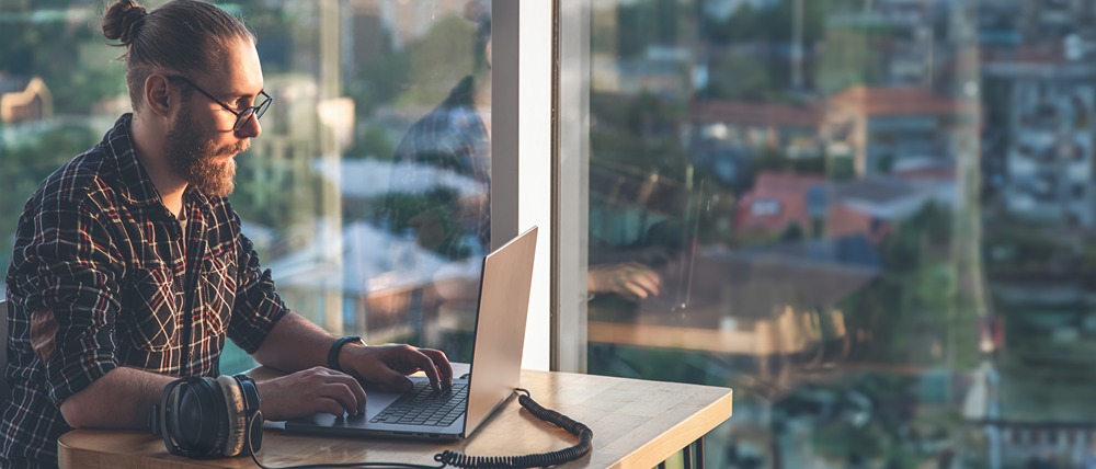 Male writing on a laptop in an high-rise office with window view of street below.