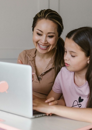 Mother smiling as her daughter works on a lap top.