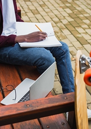 Student on a bench writing in notebook with laptop by his side.