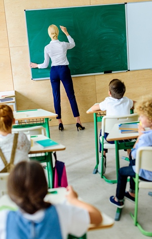 Teaching in classroom writing on green board as children watch from their desks.