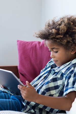 A dark skinned boy sitting on couch playing a game on a tablet.