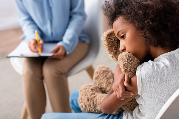 Girl in therapy session with psychiatrist sadly hugging teddy bear.