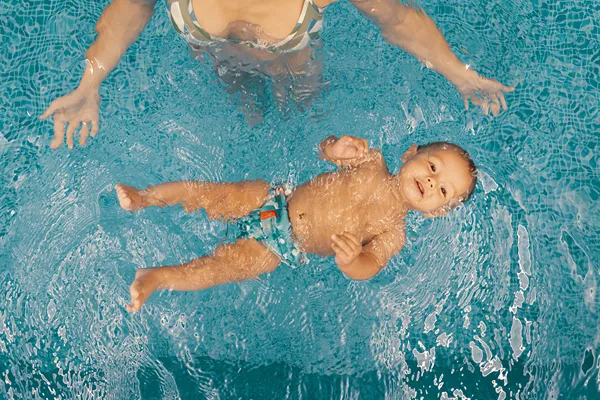 Toddler floating on his back in water and mom stands by.