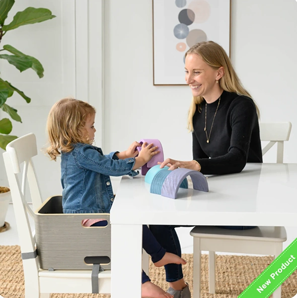 Girl sitting on booster seat on chair and playing with toys on table with her mom.