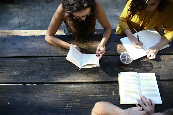 Three women outdoors on picnic table with books and notebooks.