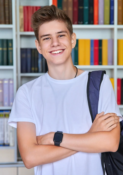 A male student with back pack stands in front of book case.