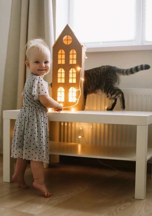 Girl playing with lit up toy house while cat explores in the background.