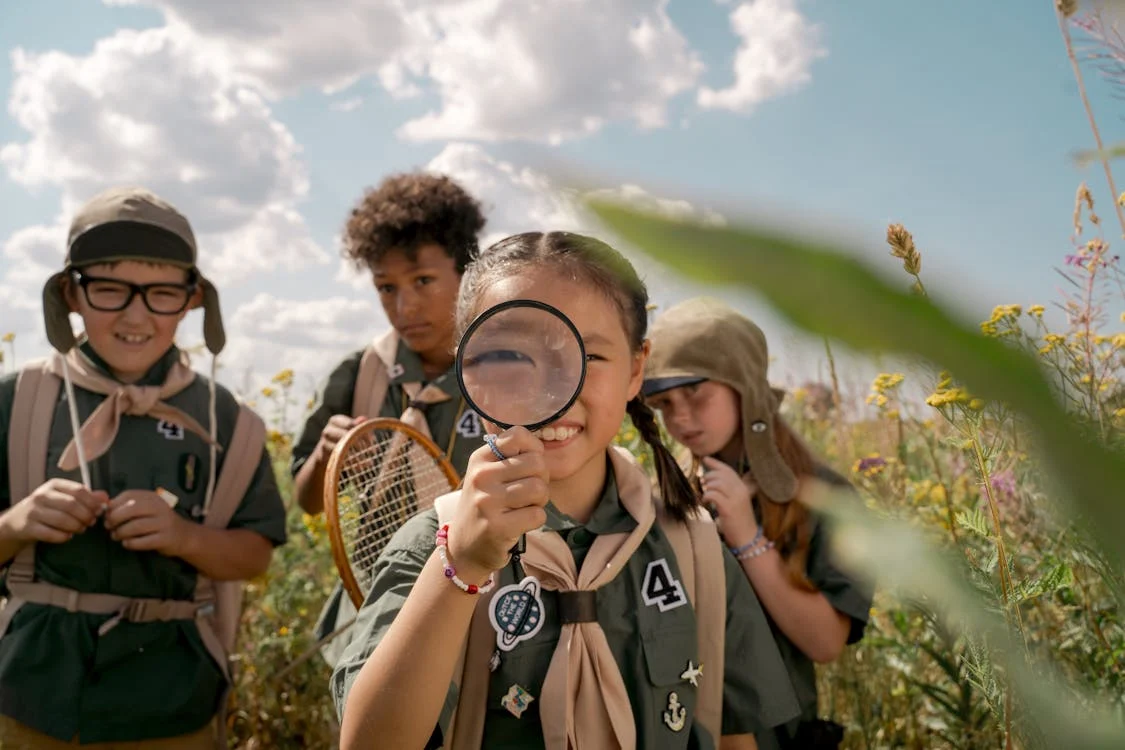 Young girl holds up magnifying glass as she explores outside with other kids.