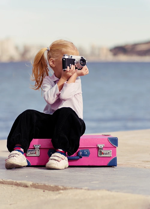 A little girl takes a picture while sitting on a suitcase on a water dock.