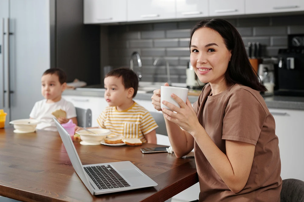 Mom at kitchen table with kids as she enjoys coffee and works on her laptop.