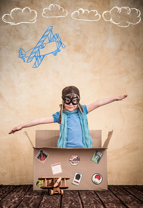 Child with goggles on spreading arms to fly in a cardboard box.