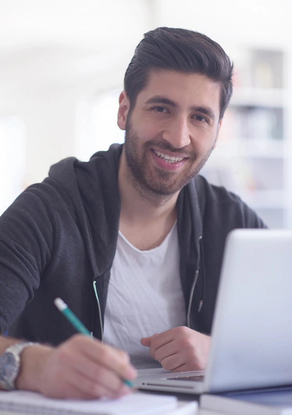 A Young adult male taking online class smiles as he looks up from his laptop .