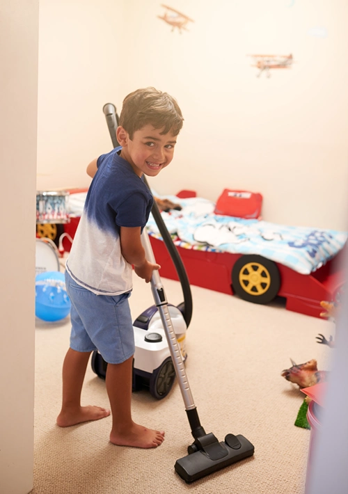 Boy looks up from vacuuming his bedroom and gives a big smile at the camera.