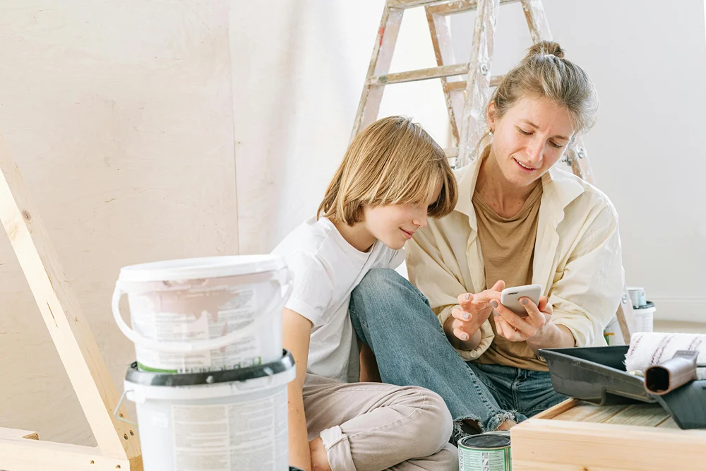 A boy surrounded by paint supplies in a room as his mom checks her phone.