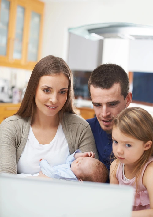 Mom holding baby using computer in the kitchen dinning room with husband and daughter.