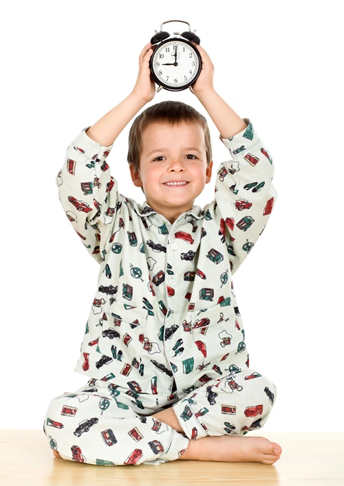 Smiling boy in pajamas holds alarm clock over his head.